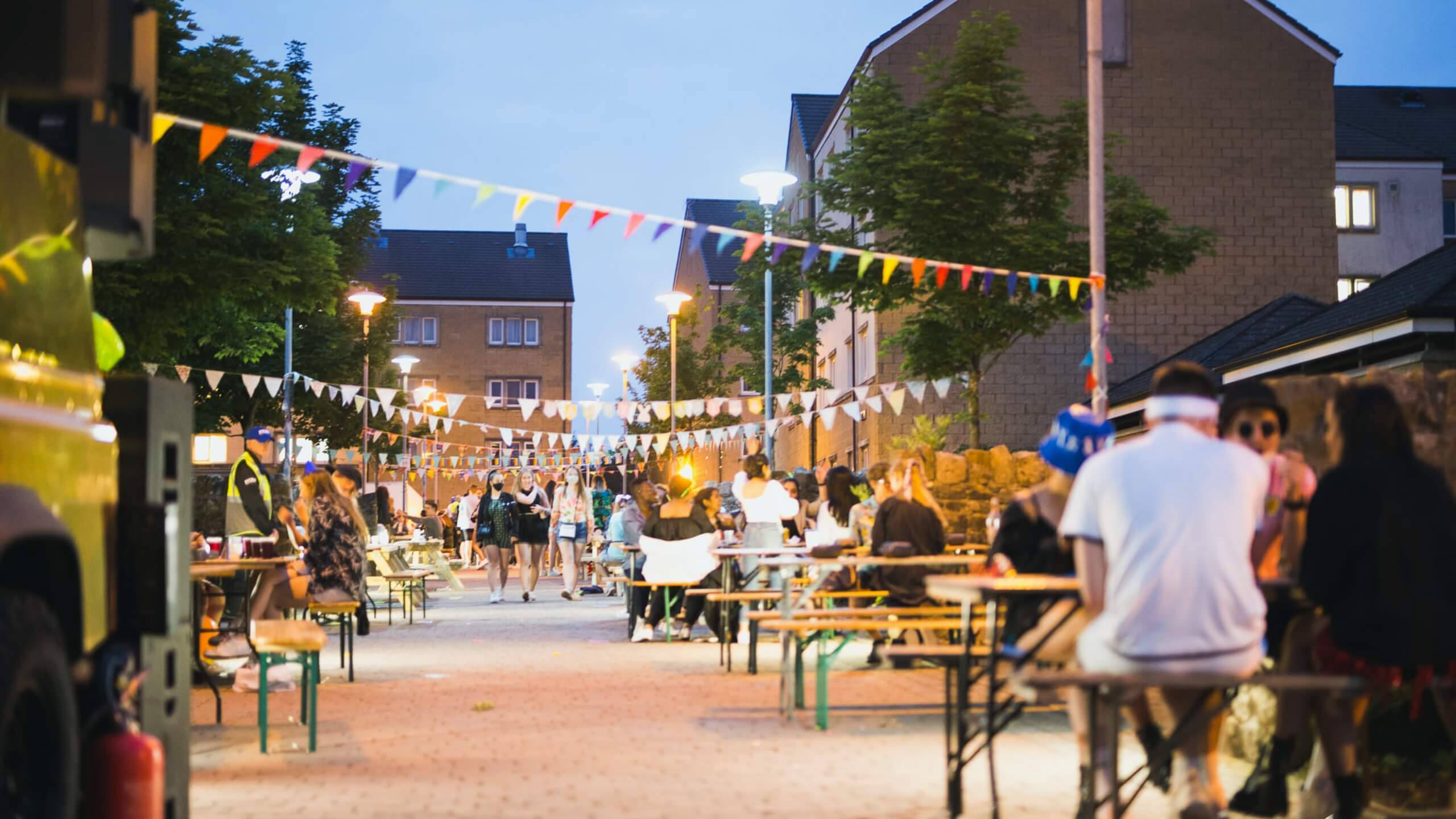 Rows of benches are filled with students in an outdoor setting at dusk. Bunting and lighting are draped across the top.