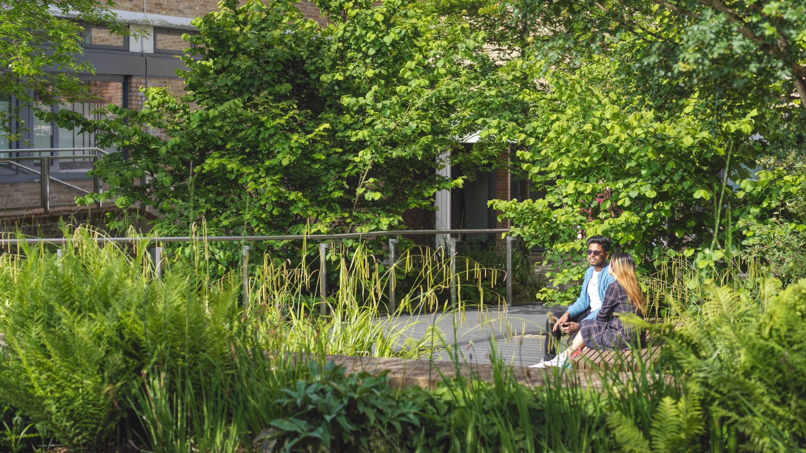 Two students sit at a bench surrounded by green foliage.